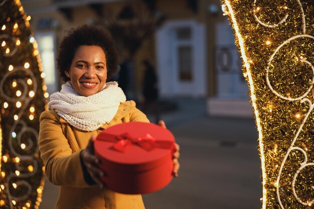 Schwarze Frau mittleren Alters in festlicher Nacht in der Stadt, die im Winter ein Geschenk auf dem Weihnachtsmarkt im Freien hält.