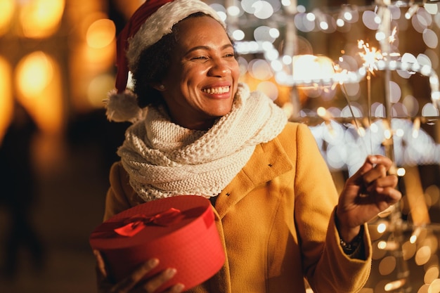 Schwarze Frau mittleren Alters mit Wunderkerzen und Weihnachtsgeschenkbox in festlicher Winternacht in der Stadt.