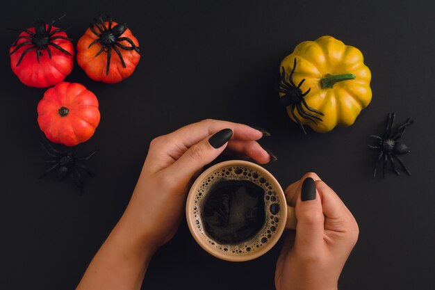 Schwarze, gepflegte Hände, die Cappuccino-Becher auf Halloween-Hintergrund mit hingebungsvollen Kürbissen und Spinnen halten. Sicht von oben