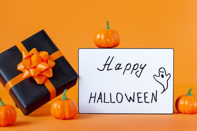 Schwarze Geschenkbox mit orangefarbenem Band und Kürbis und Leuchtkasten mit dem Satz Happy Halloween auf orangem Hintergrund.