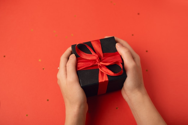Schwarze Geschenkbox mit rotem Band in den Händen der Frau auf Rot. Valentinstag Konzept.