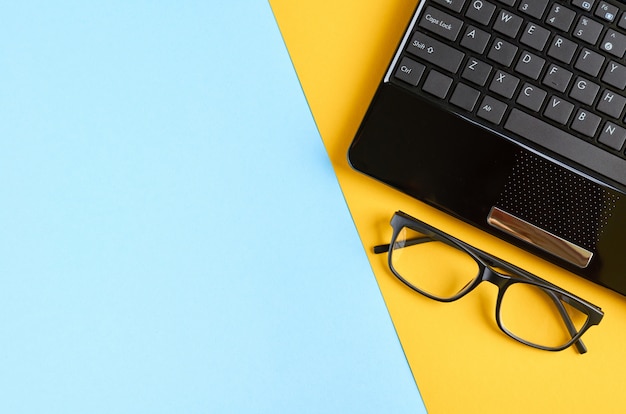 Schwarze Gläser und Laptoptastatur auf blauer und gelber Oberfläche.