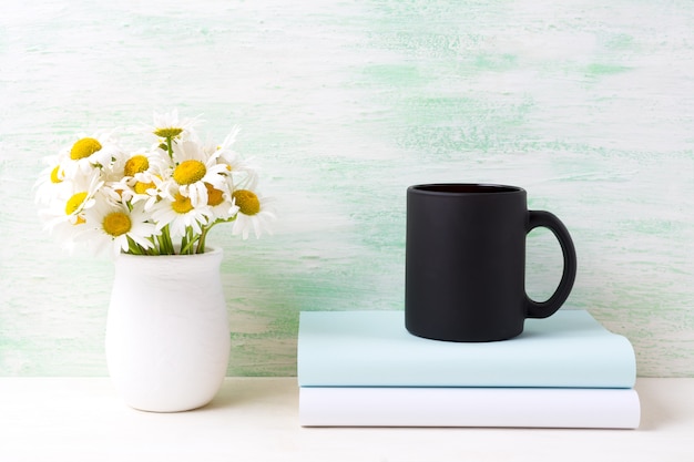 Schwarze Kaffeetasse mit weißem Feldkamille-Bouquet in handgemachter rustikaler Vase und Büchern