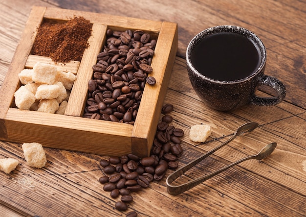 Schwarze Keramikschale des frischen rohen Bio-Kaffees mit Bohnen und gemahlenem Pulver mit Rohrzucker in der Weinleseschachtel auf hölzernem Hintergrund.