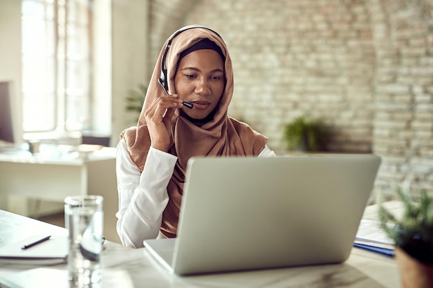 Schwarze muslimische Kundendienstmitarbeiterin spricht mit ihrem Kunden, während sie im Callcenter am Laptop arbeitet