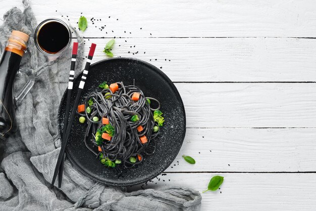 Schwarze Pasta mit Gemüse auf einem schwarzen Teller Draufsicht auf weißem Holzhintergrund Freier Kopierplatz