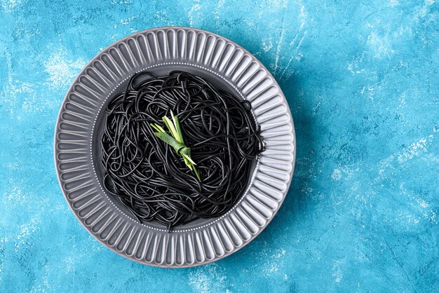 Schwarze Spaghetti der Draufsicht mit Tintenfisch-Tinte auf grauer Platte mit grünem Blatt auf blauem Hintergrund mit Kopienraum