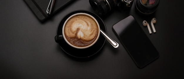 Schwarze Tasse Kaffee auf dunklem Schreibtisch mit Kamera, Smartphone, Zeitplanbuch und kabellosem Kopfhörer