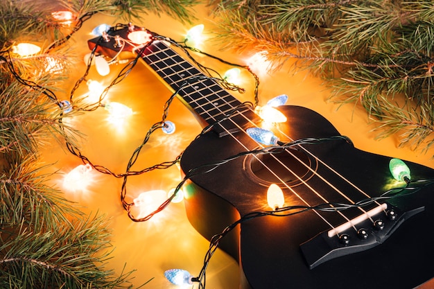 Schwarze Ukulele-Gitarre und Weihnachtsschmuck auf dunklem Hintergrund. Hawaiianische Gitarre mit beleuchteter Girlande auf dunklem Hintergrund. Klassische Formen der Geschenkgitarre für Weihnachten, neues Jahr.