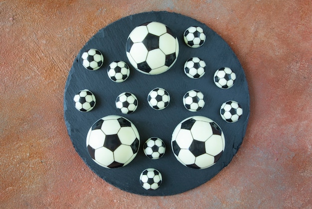 Schwarze und weiße klassische Fußballbälle auf einer schwarzen runden Servierplatte - Dessertkuchen aus schwarzer und weißer Milchschokolade auf dem Feldhintergrund