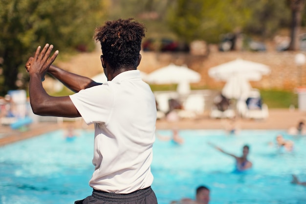 Schwarzer athletischer Hotel-Resort-Animator, der vor dem Pool trainiert, Form hinter verschwommenem Menschenhintergrund - Wassergymnastik-Aktivität