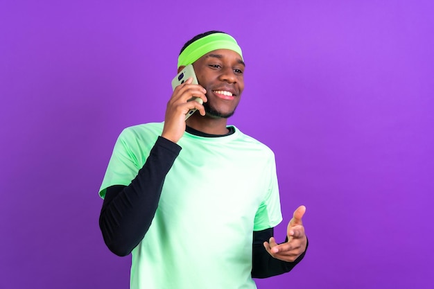 Schwarzer ethnischer Mann mit einem Telefon in grüner Kleidung isoliert vor violettem Hintergrund, der lächelnd am Telefon spricht