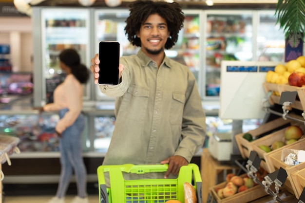 Schwarzer Kerl zeigt Smartphone mit leerem Bildschirm beim Einkaufen von Lebensmitteln im Supermarkt