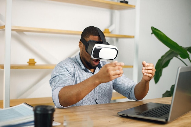 Schwarzer männlicher Unternehmer, der im Büro sitzt und eine Virtual-Reality-Brille verwendet