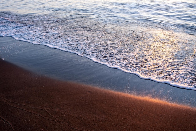 Schwarzer Magnetit-Sand und Meereswellen unter Sonnenuntergang strahlen Kupfer-Highlights auf Sand verschwommenen Meereslandschaftshintergrund Foto in hoher Qualität