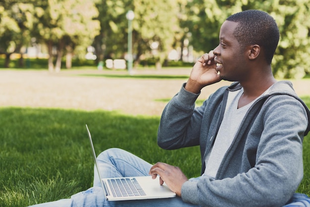Schwarzer Mann, der draußen auf Gras mit Laptop sitzt und auf Smartphone spricht, Seitenansicht, Kopienraum