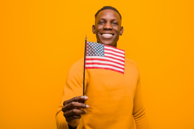 Schwarzer Mann des jungen Afroamerikaners gegen die orange Wand, die eine USA-Flagge hält