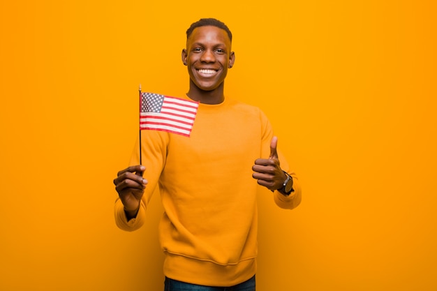 Schwarzer Mann des jungen Afroamerikaners gegen die orange Wand, die eine USA-Flagge hält