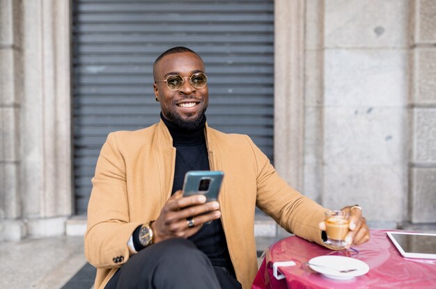 Schwarzer Mann sitzt in einem Tischcafé, trinkt Kaffee, während er das Smartphone benutzt und lächelt in die Kamera schaut.