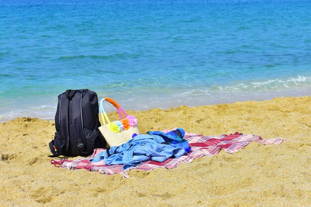 Foto schwarzer rucksack, gewebte tasche und rosa plaid an einem sandstrand und ruhigem blauen meer