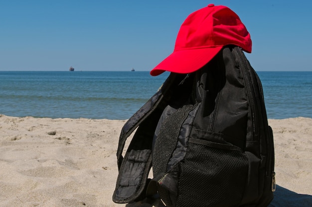 Schwarzer Rucksack mit zwei Riemen und roter Kappe darauf am grauen Sandstrand des ruhigen blauen Meeres