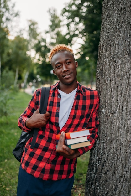 Schwarzer Student mit Buch und Rucksack, der im Sommerpark nahe dem Baum steht. Ein Teenager, der im Freien lernt und zu Mittag isst