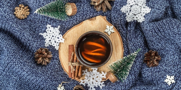 Schwarzer Tee in Keramiktasse mit traditioneller Neujahrsdekoration. Kleine Tannen, gehäkelte Schneeflocken, Zimt und Zapfen. Holzständer, kuscheliges Strickpullover-Arrangement, Bannerformat