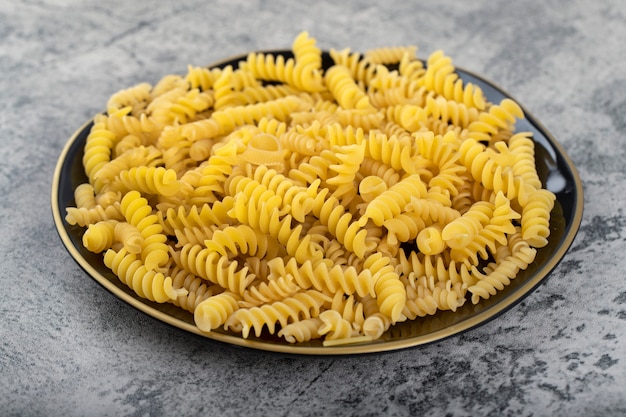 Schwarzer Teller mit rohen Fusilli-Nudeln auf Steintisch.