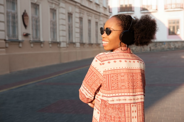Schwarzes afroamerikanisches Mädchen hat einen Spaziergang in der Abendstadt und hört Musik in Kopfhörern. Mädchen in der Sonnenbrille im Sommer lächelt und schaut auf Sonnenuntergang.