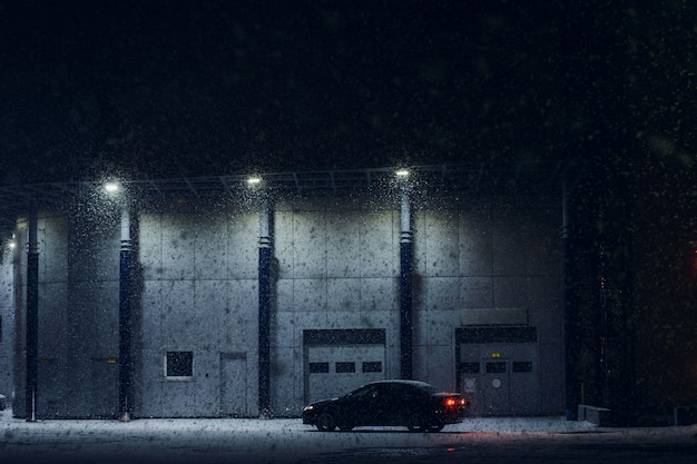 Schwarzes Auto geparkt nahe Bürogebäude im Winterabend im starken Schneesturm. Auto in der Nähe von Auto-Service in der Nacht. Maschine in der Nähe des automatischen Tors zur Wartung. Moderne Architektur in dunkler Nacht.