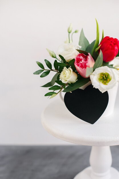 Schwarzes Brett Herz Exemplar. Frühlingsblumenstrauß in weißer Vase auf weißem Holzständer. Rosen, Tulpen und Lisianthus.