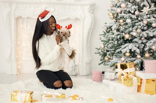 Schwarzes Mädchen mit Weihnachtsmütze, das in der Nähe des Weihnachtsbaums sitzt und einen kleinen Hund hält