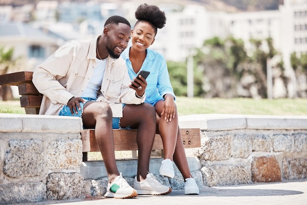Schwarzes Paar liebt und telefoniert, während es im Internet in sozialen Medien surft oder ein Video ansieht, während es auf einer Parkbank sitzt Glücklicher Mann und Frau auf einem romantischen Date-Trip oder Sommerurlaub, um sich gemeinsam zu entspannen
