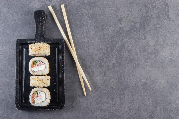 Schwarzes Schneidebrett von Sushirollen mit Sesam auf Steinhintergrund.