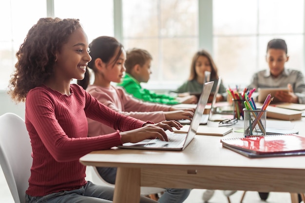 Schwarzes Schulmädchen mit Laptop, das mit multiethnischen Klassenkameraden im Klassenzimmer sitzt