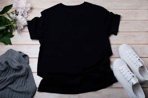 Schwarzes T-Shirt-Modell aus Baumwolle für Damen mit grauem Aran-Pullover, weißen Turnschuhen und blassrosa Pfingstrose. Design-T-Shirt-Vorlage, T-Shirt-Druck-Präsentationsmodell