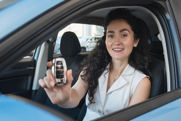 Schwarzhaarige Frau zeigt Autoschlüssel, die am Lenkrad eines blauen Automobils sitzen