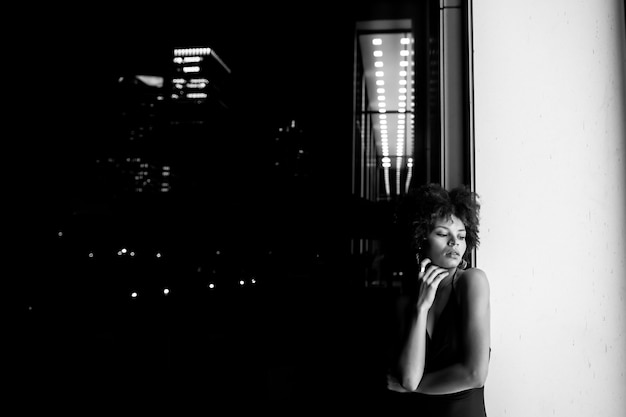 Schwarzweiss-Foto der jungen afroamerikanischen schwarzen Frau bei Nacht. Frau, die nahe dem beleuchteten Fenster auf der Straße aufwirft. Nachtleben, Mode und Stilkonzept. Platz für Text
