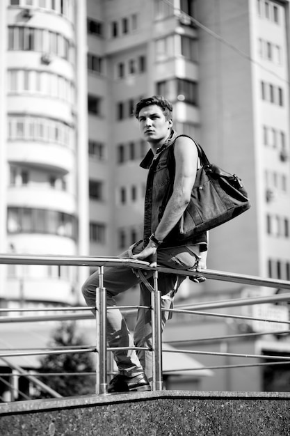 Schwarzweißfoto eines Modetyps mit einer Tasche, der auf dem Hintergrund der Stadt sitzt
