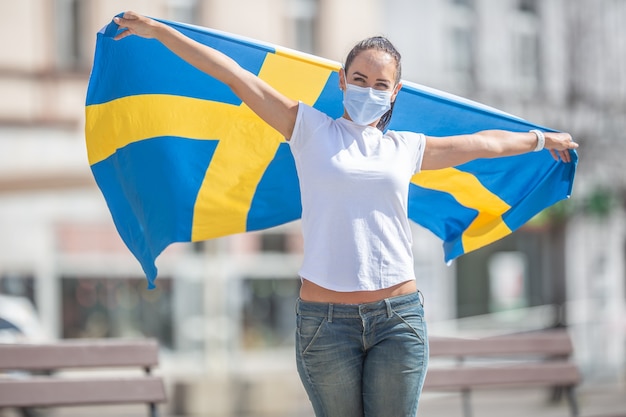 Schwedisches Mädchen auf einer Straße hält die Flagge in den Händen und feiert, trägt eine Gesichtsmaske.