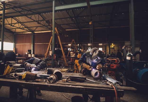 Schweißer in Schutzkleidung und Maske schweißen Metallrohr auf dem Industrietisch, während Funken fliegen.