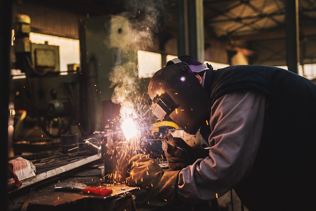 Schweißer in Schutzkleidung und Maske schweißen Metallrohr auf dem Industrietisch, während Funken fliegen.