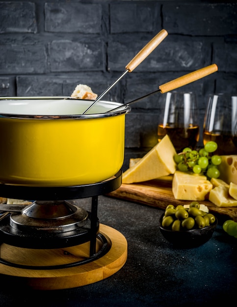 Schweizer Feinschmeckerfondue im traditionellen Fonduetopf mit Gabeln, verschiedenen Käsesorten, Oliven, Brot und Trauben