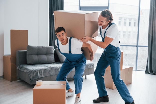 Schwere Kisten Zwei junge Mover in blauer Uniform, die drinnen im Raum arbeiten