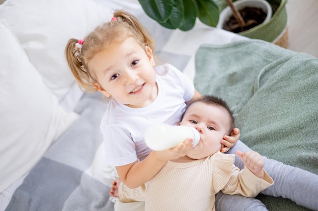 Schwestern füttern zu Hause Babymilch aus einer Flasche auf dem Bett Die ältere Schwester hält das Baby zu Hause auf dem Bett das Konzept der Babynahrung