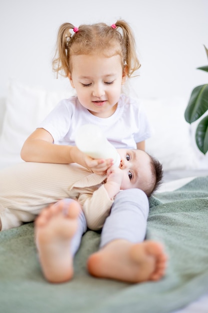 Schwestern füttern zu Hause Babymilch aus einer Flasche auf dem Bett Die ältere Schwester hält das Baby zu Hause auf dem Bett das Konzept der Babynahrung