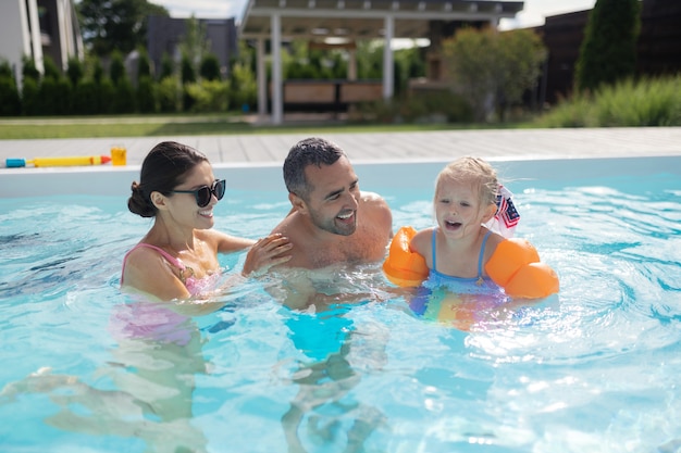 Schwimmen mit den Eltern. Süßes blondes Mädchen, das sich beim Schwimmen mit den Eltern aufgeregt und erstaunlich fühlt