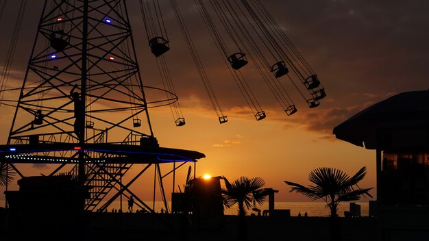 Schwingende Karussell-Kreisverkehr-Kettenfahrt bei Sonnenuntergang. Unterhaltung am Strand, Silhouetten von Palmen vor dem Hintergrund des Sonnenuntergangs am Meer