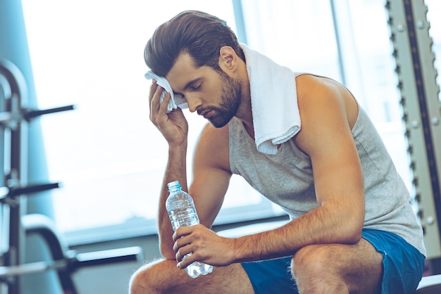 Schwitzend nach tollem Training. Hübsche junge Männer in Sportkleidung peitschen Schweiß mit seinem Handtuch und halten eine Wasserflasche, während sie im Fitnessstudio sitzen