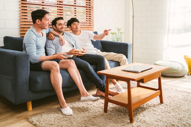 Schwule Menschengruppe sitzt auf dem Sofa mit Popcorn und genießt es, Inhalte auf dem Fernseher im Wohnzimmer anzusehen Drei LGBT-kaukasische und asiatische Menschen leben zusammen Vielfalt der schwulen Freundschaft